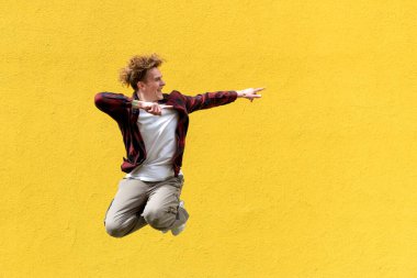 young guy student jumps against the background of yellow isolated wall and shows his hands to the side on copy space, energetic male dancer flies in the air and advertises an empty space clipart