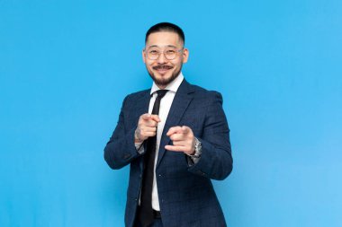 Hey sen! Takım elbiseli ve gözlüklü başarılı Asyalı işadamı sizi mavi izole edilmiş arka planda seçiyor, iş elbiseli Koreli girişimci davet ediyor ve karşılıyor