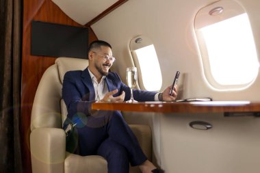 Takım elbiseli Asyalı işadamı ve şampanya bardaklı bardaklar lüks jette uçuyor ve akıllı telefon, Koreli girişimci filmlerini telefonla kullanıyor ve video aracılığıyla iletişim kuruyor.