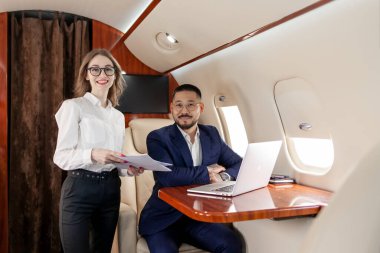 İş adamları özel uçakla uçuyor, başarılı Asyalı iş adamı takım elbiseli, elinde laptop ve belgelerle kadın asistan uçuyor ve kameraya bakıyorlar.