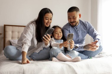 Asyalı küçük kız ailesiyle yatakta uzanıyor ve akıllı telefon kullanıyor. Anne ve babası küçük kızına cep telefonu kullanmayı öğretiyor. Cep telefonlarıyla mutlu Koreli bir aile.
