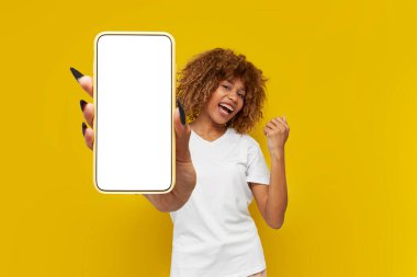 Mutlu kıvırcık Amerikalı kız kazananı boş akıllı telefon ekranı gösteriyor ve sarı izole arka planda zaferini kutluyor, genç kadın telefon ekranında fotokopi alanı reklamları yapıyor ve şanslı bir şekilde seviniyor.