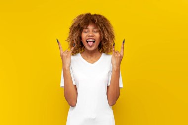 Beyaz tişörtlü çılgın kıvırcık Amerikalı kız dil ve rock jesti gösteriyor. Sarı yalıtılmış arka planda elleri olan, saç stili ve diş telli komik kadın ağır müzik sembolü gösteriyor.