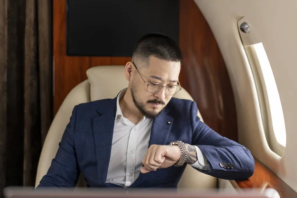 successful asian businessman in suit and glasses flies in private luxury plane and looks at his watch, korean trader financier is late and rushes to meeting in plane, luxury lifestyle