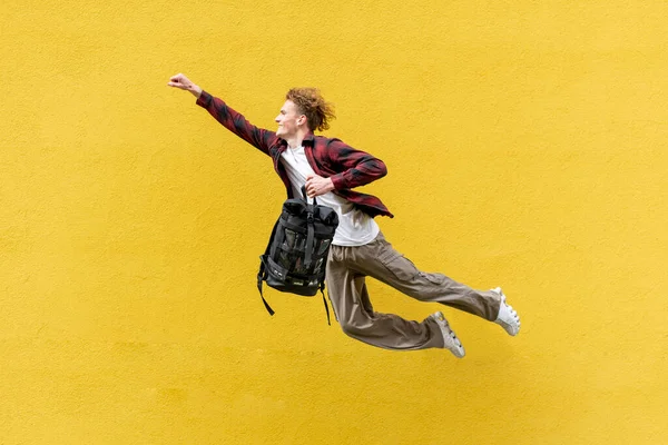 バックパックを持つ若い男学生は前方に飛ぶと研究するために急いで スーパーマンの男はジャンプをポーズし 黄色の隔離された壁に対して空気中で実行されます 休暇や旅行の概念 — ストック写真