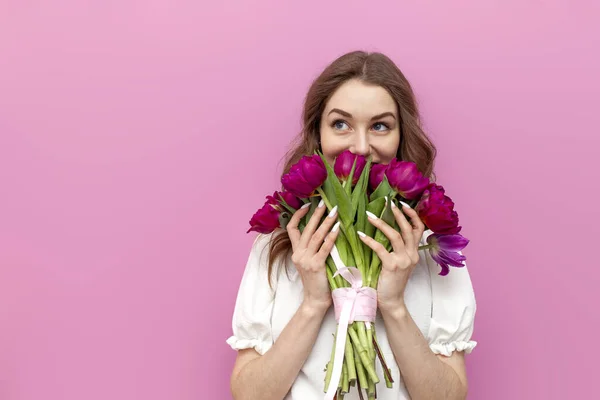 Stock image cute young woman in festive outfit holds bouquet of pink tulips and smells them on pink isolated background, girl with flowers for the holiday of March 8