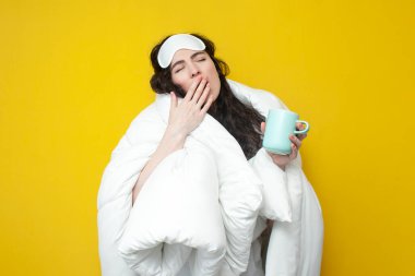 Uyku maskesiyle örtülü uykulu kız elinde bir fincan kahve, sarı arka planda esniyor yorgun kadın uyumak ve içmek istiyor.