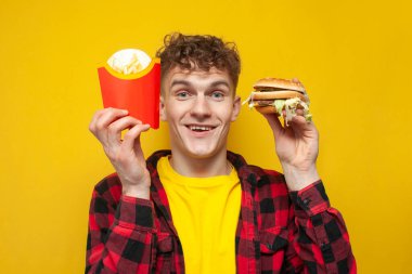 Genç ve çekici bir öğrenci elinde fast food tutuyor ve sarı arka planda sürpriz yapıyor. Hamburger ve patates kızartmalı adam.