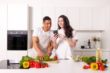 multiracial couple in kitchen preparing salad together and looking at smartphone, american guy and european girl preparing veggie food and looking at phone in kitchen, happy young interracial family