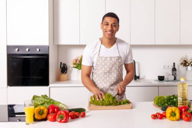 Önlüklü genç Afrikalı Amerikalı adam beyaz modern mutfakta yeşillik kesiyor, aşçı evde sebze ve bitki salatası hazırlıyor.