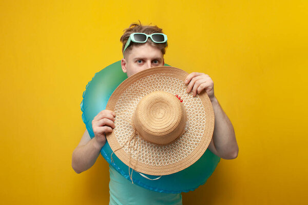 молодой парень летом на отдыхе с незамысловатым купальным кругом стесняется и прячется за большой соломенной шляпой на желтом фоне