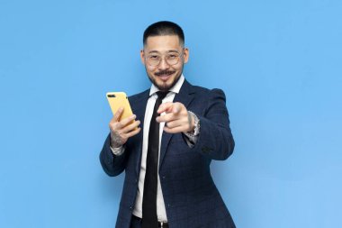 Hey sen! Takım elbiseli ve gözlüklü başarılı Asyalı işadamı akıllı telefon kullanır ve sizi mavi izole edilmiş arka planda seçer, iş elbiseli Koreli girişimci telefon davetleriyle ve hoş geldin mesajlarıyla