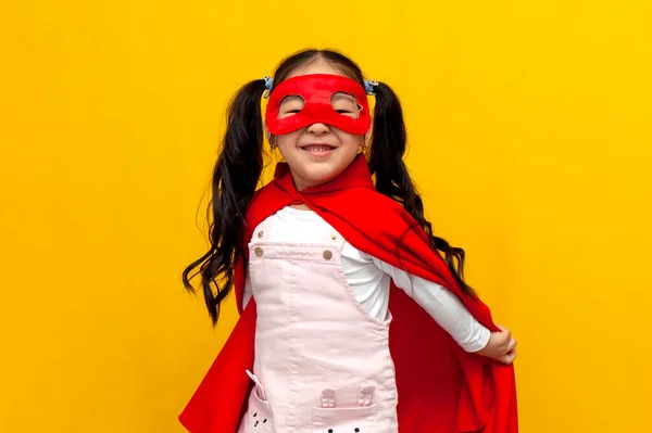 little asian girl in superman costume and mask smiles, korean child in superhero cape on yellow isolated background
