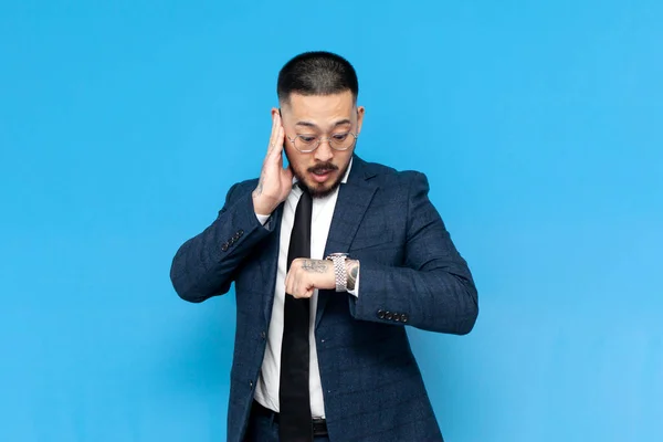 stock image successful asian businessman in suit and glasses is late and looks at his watch on blue isolated background, korean entrepreneur in business clothes checks the time