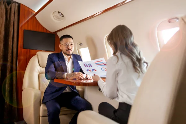 business people at meeting in private plane, asian businessman in suit discusses work with female colleague, manager shows graphs and reports to the boss, business partners fly in jet