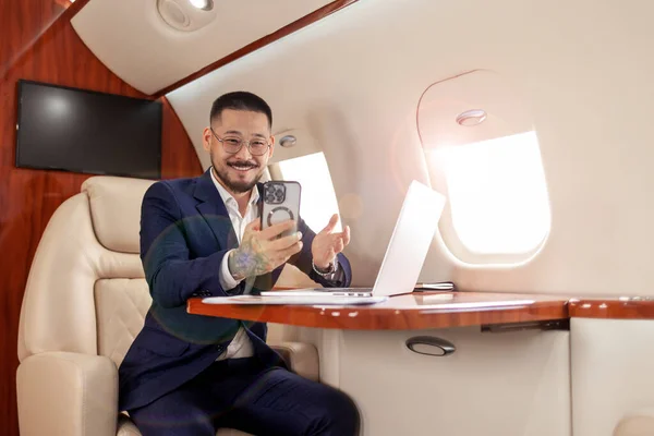 young asian businessman is sitting in private luxury jet with laptop and chatting via smartphone video call, korean manager financier in suit and glasses talking on the phone online video broadcast