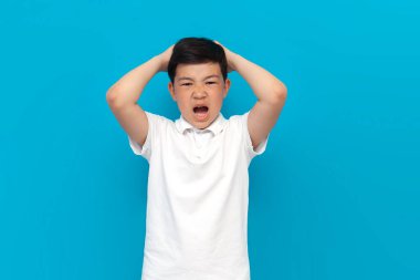 Beyaz tişörtlü küçük Asyalı çocuk bağırıyor ve kafasını mavi arka planda tutuyor. Koreli çocuk stresli ve depresyonda.