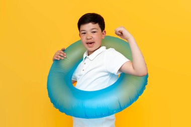 Küçük Asyalı çocuk şişme yüzme yüzüğü tutuyor ve sarı arka planda zaferi kutluyor. Koreli çocuk yaz tatilinde denize gidiyor ve başarıyla ve iyi şansla seviniyor.