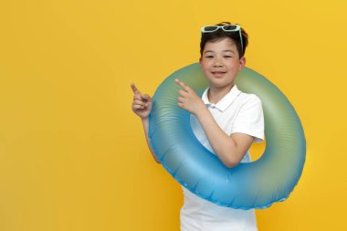 Tatildeki küçük Asyalı çocuk şişme yüzme yüzüğünü tutuyor ve ellerini sarı arka planda yana uzatıyor. Koreli çocuk denize gidiyor ve uzayın fotokopisini çekiyor.