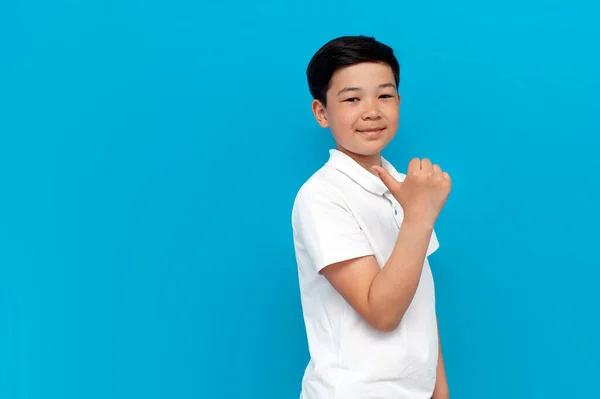 little asian boy in white t-shirt points back on blue background, korean child advertises copy space from behind on isolated background