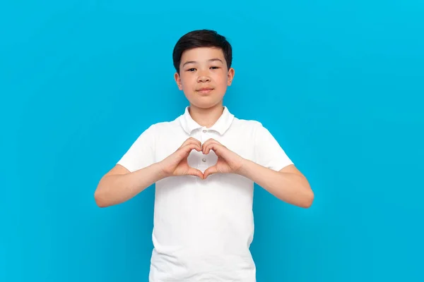 stock image little asian boy in white t-shirt shows heart with his hands on blue background, korean child shows gesture of love and romance on isolated background