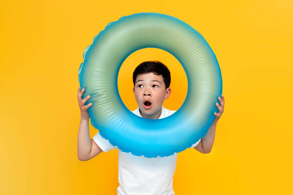 маленький азиатский мальчик с надувным плавательным кольцом и удивленно глядя на желтый фон, пораженный корейский ребенок отправляется в отпуск на море летом