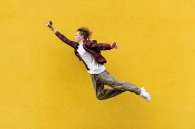 Sırt çantası ve kahve taşıyan genç bir öğrenci hızla ilerler ve çalışmak için acele eder, enerjik bir adam sarı izole edilmiş duvara, tatile ve seyahat konseptine karşı havaya atlar ve koşar.