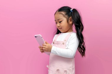 Pembe elbiseli küçük Asyalı kız akıllı telefon kullanıyor ve pembe izole edilmiş arka planda interneti seçiyor, Koreli anaokulu çocuğu telefonu tutuyor ve ekranda tıklıyor.