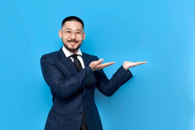 Takım elbiseli ve gözlüklü başarılı Asyalı işadamı ellerini yana doğru gösteriyor, iş kıyafetlerindeki Koreli girişimci noktaları ve reklamlar alanı kopyalıyor