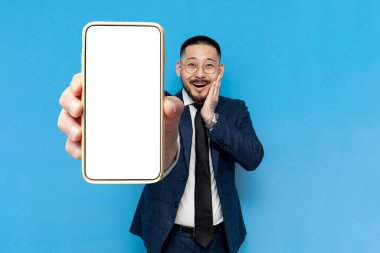 Takım elbiseli ve gözlüklü başarılı Asyalı iş adamı mavi izole edilmiş arka planda boş bir akıllı telefon ekranı, Koreli iş adamı telefon ekranı sunar ve şaşırır.