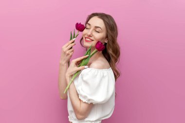 Genç kız yüzünün tenine iki pembe lale ile dokunuyor ve pembe izole edilmiş arka planda gülümsüyor. Beyaz elbiseli, çiçekli pozlar veren kadın.