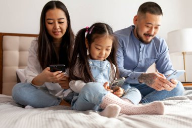 Mutlu, genç Asyalı bir aile yataklarında akıllı telefonlar kullanıyor. Koreli bir baba, anne ve küçük bir kız telefona bakıyor ve mesaj yazıyor.