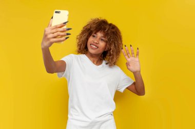 Genç kıvırcık saçlı Amerikalı kız akıllı telefon selamları kullanıyor ve sarı izole edilmiş arka planda avuç içi gösteriyor. Beyaz tişörtlü Afrikalı kadın video bağlantısıyla iletişim kuruyor ve hoşça kal diyor.