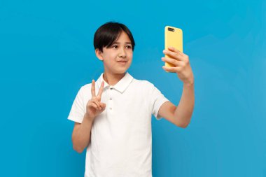 Beyaz tişörtlü Asyalı çocuk kendini akıllı telefonuyla çekiyor ve mavi izole edilmiş arka planda barış jesti gösteriyor, Koreli çocuk selfie çekiyor ve video aracılığıyla iletişim kuruyor.