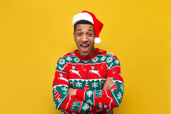 stock image african american guy in New Year's clothes and santa hat is surprised and shouts on yellow isolated background, amazed man with crossed arms and open mouth