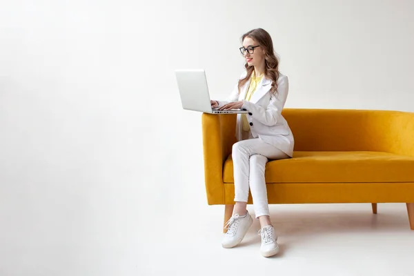 若いビジネスマンスーツの女性は快適な柔らかいソファに座って ラップトップを使用して 正式な摩耗の女の子は白い隔離された背景に黄色のソファの上にコンピュータに入力されています — ストック写真