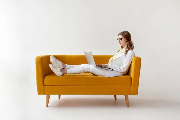 スーツ姿の若い実業家が快適なソファーでラップトップを使用しています 正式に身に着けている女の子は白い隔離された背景に黄色のソファの上にコンピュータに入力されています — ストック写真