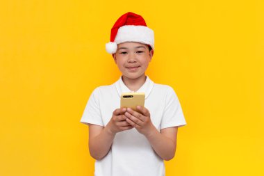 Noel Baba şapkalı küçük Asyalı çocuk sarı izole edilmiş arka planda akıllı telefon kullanıyor. Yılbaşı için internette Koreli çocuk şapkası takıyor.