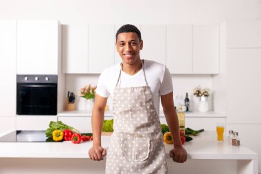 Önlüklü genç Afrikalı Amerikalı adam beyaz modern mutfakta duruyor ve gülümsüyor. Adam aşçı ve ev hanımı sebze ve otlarla dolu bir masanın yanında duruyor.