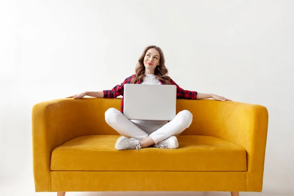 若いかわいい女の子は柔らかい快適なソファと夢でラップトップを使用しています 高価な女性はコンピュータ上でオンラインで入力し 白い隔離された背景に黄色のソファに考えています — ストック写真