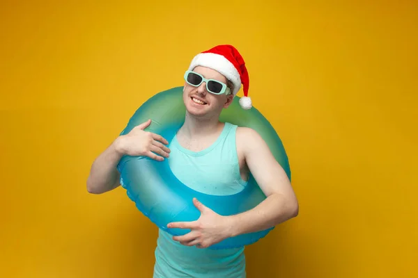 young guy in a New Year's hat in the summer on vacation with an inflatable swim ring rejoices and smiles, a man with a swim ring for Christmas
