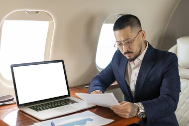 Takım elbiseli ve gözlüklü başarılı Asyalı işadamı özel bir jette oturuyor ve boş ekranlı dizüstü bilgisayarın yanında belgelerle çalışıyor. Koreli finansör uçağa biniyor ve evraklara bakıyor, boşluğu kopyalıyor.