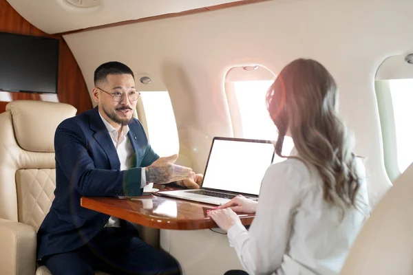 business people at meeting in private plane, Asian businessman in suit is talking to woman in jet and pointing at blank laptop screen, colleagues are discussing deal and flying in plane