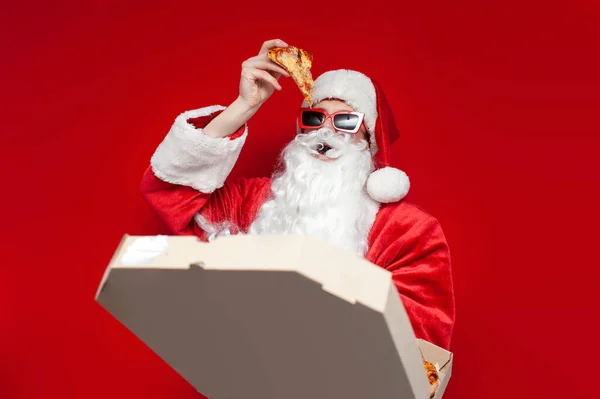 サンタ クラス グラスはピザの箱を背景色に帽子の男とクリスマスのためのサンタの衣装はファーストフードを食べる — ストック写真