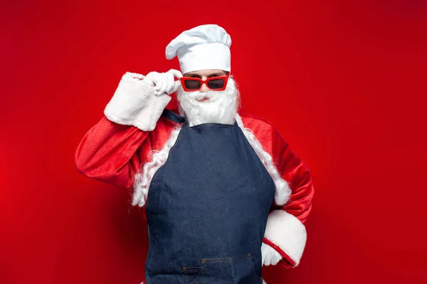 シェフの帽子のサンタ クラスと赤い背景のエプロンサンタの衣装の男性料理人は眼鏡をかけクリスマスにカメラを見る — ストック写真
