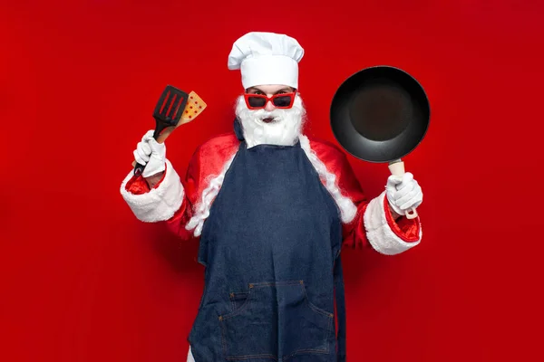 Weihnachtsmann Kochmütze Und Kochschürze Hält Pfanne Und Utensilien Auf Rotem — Stockfoto