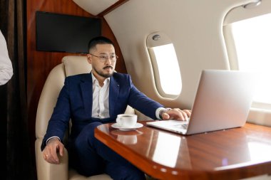Takım elbiseli ve gözlüklü başarılı Asyalı işadamı özel lüks bir uçakta dizüstü bilgisayarla uçuyor, Koreli tüccar finansörü uçakta bilgisayar işinde çalışıyor, lüks bir yaşam tarzı.