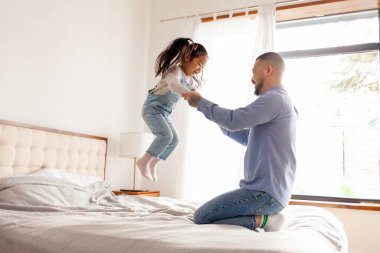 Küçük, aktif bir Asyalı kız babasının yatağında zıplıyor ve dans ediyor. Koreli bir adam kızıyla oynuyor ve elini tutuyor.