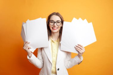 Beyaz takım elbiseli genç bir iş kadını elinde turuncu izole edilmiş arka planla ilgili belgeler tutuyordu. Kâğıtlı gözlüklü kız müdürü, son teslim tarihi konsepti, ofis çalışanı tarzı.