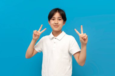Beyaz tişörtlü, Asyalı bir çocuk elleriyle barışı işaret ediyor ve mavi izole edilmiş arka planda gülümsüyor. Koreli bir çocuk.